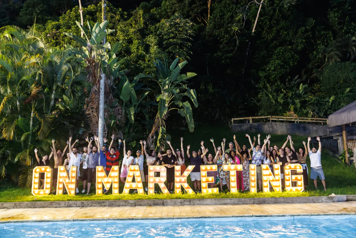 Time da On comemorando com os braços para o alto atrás do letreiro luminoso ON MARKETING, à beira da piscina